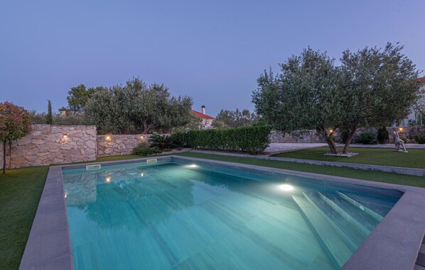 A rectangular swimming pool with a stone wall on one side and a grassy area with trees and a statue on the other. - Amazing home in Zaton (Zaton)