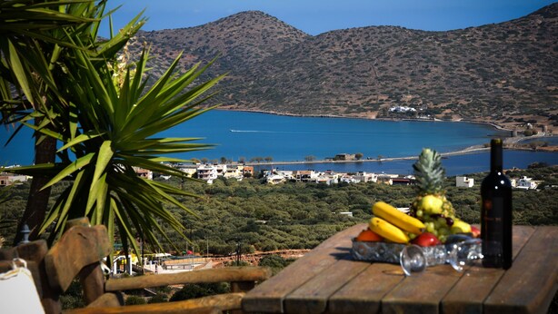 A table with fruits, a bottle, and glasses, overlooking a coastal area with mountains and buildings. - Elounda Theo Villa (Agios Nikolaos)