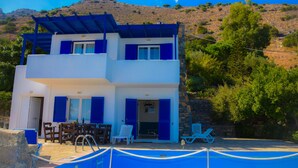 A two-story building with a pool area, outdoor seating, and a mountainous backdrop. - Elounda Theo Villa (Agios Nikolaos)