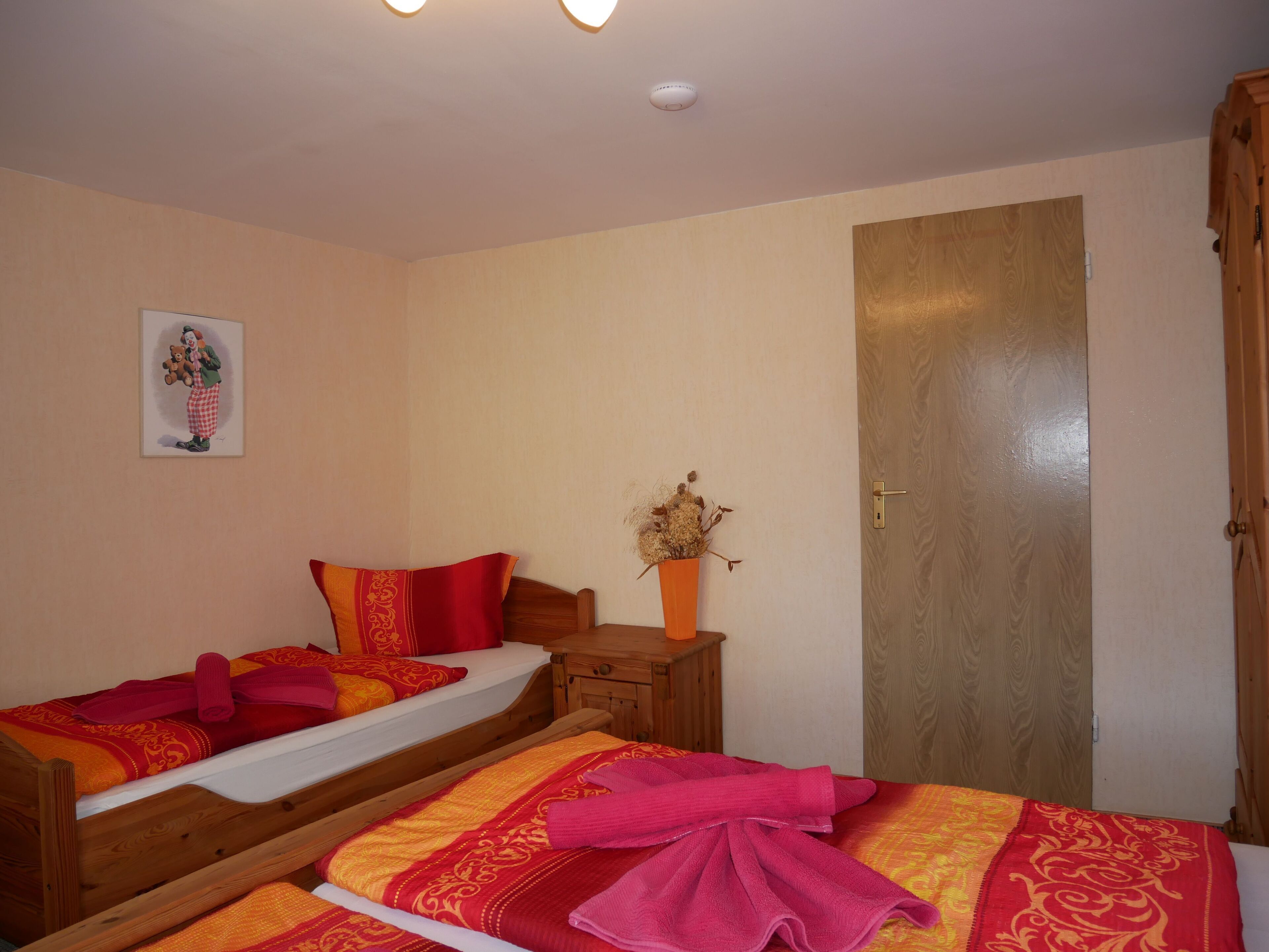 A room with two wooden beds, each with red and yellow bedding, a wooden nightstand with a vase of dried flowers, and a door with a golden handle.