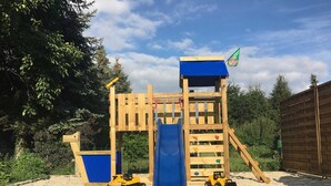 A wooden play structure with a slide and climbing area, surrounded by a stone-tiled patio and greenery.