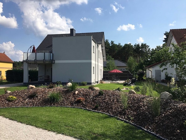 Ein modernes, zweistöckiges Haus mit Balkon, umgeben von einem gepflegten Garten mit Steinen und Sträuchern.