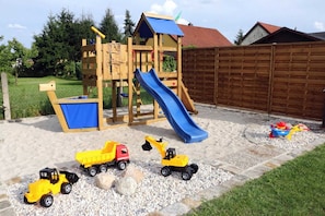 Eine hölzerne Spielkonstruktion mit Rutsche und Kletterelementen, umgeben von einem Kiesplatz mit Spielzeugfahrzeugen.