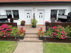 A white building with a red-tiled roof, two white doors, and a brick pathway leading to a wooden bench area with potted plants.