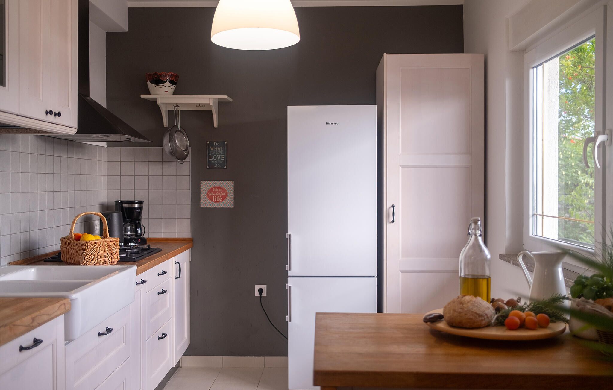 Une cuisine moderne avec des meubles blancs, un réfrigérateur blanc, un plan de travail en bois et une fenêtre donnant sur un paysage verdoyant.