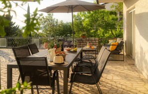 Patio esterno con tavolo apparecchiato per mangiare, sedie, ombrellone e muretto in pietra con vegetazione sullo sfondo.