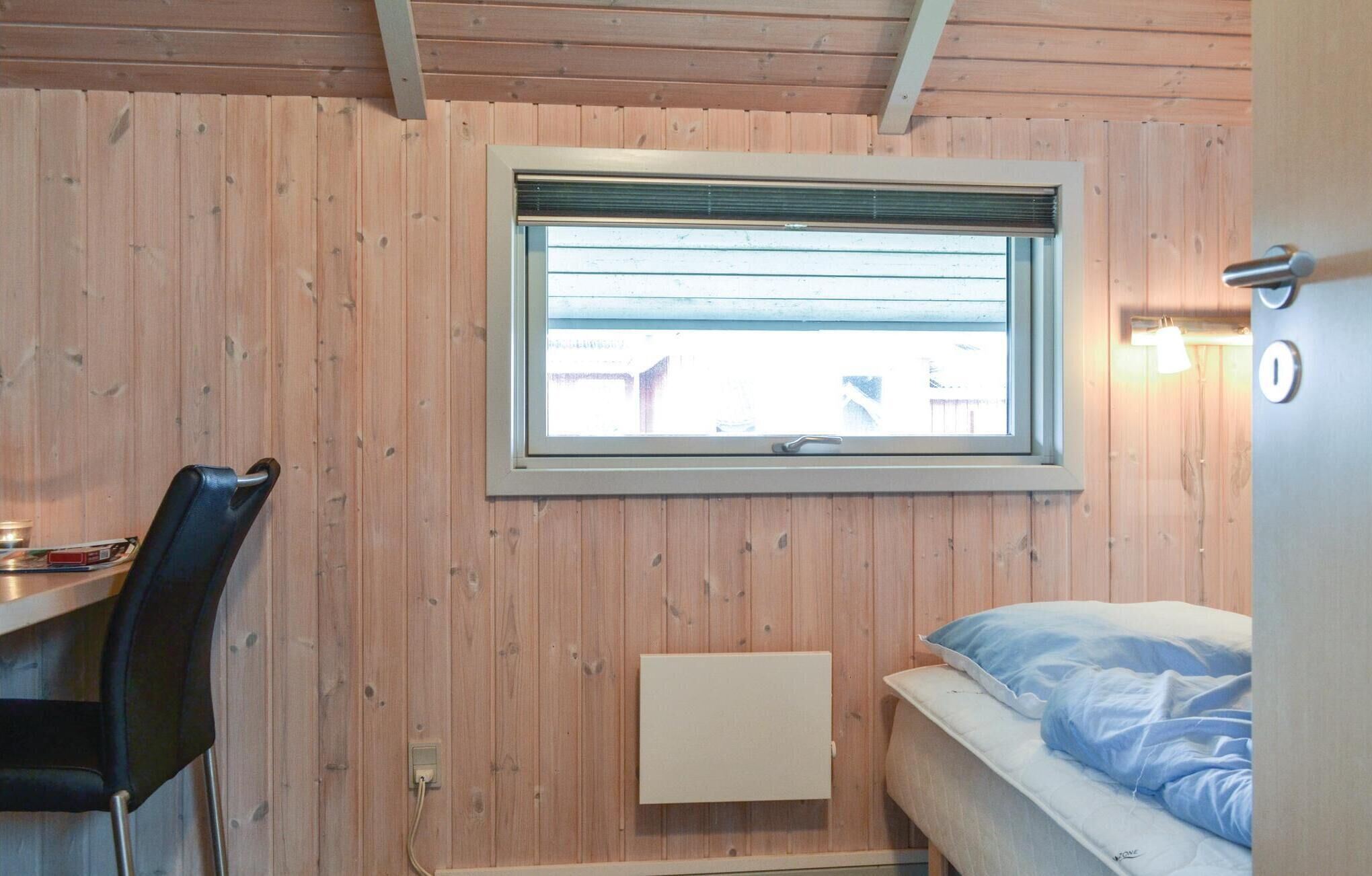 Ett litet rum med en enkel säng, ett skrivbord med en stol, ett fönster med persienner och en väggmonterad lampa.
