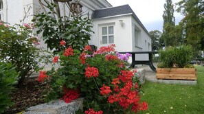Une maison blanche avec un toit noir, entourée d’un jardin où poussent des fleurs rouges et roses, avec un banc en bois et une pelouse verdoyante.