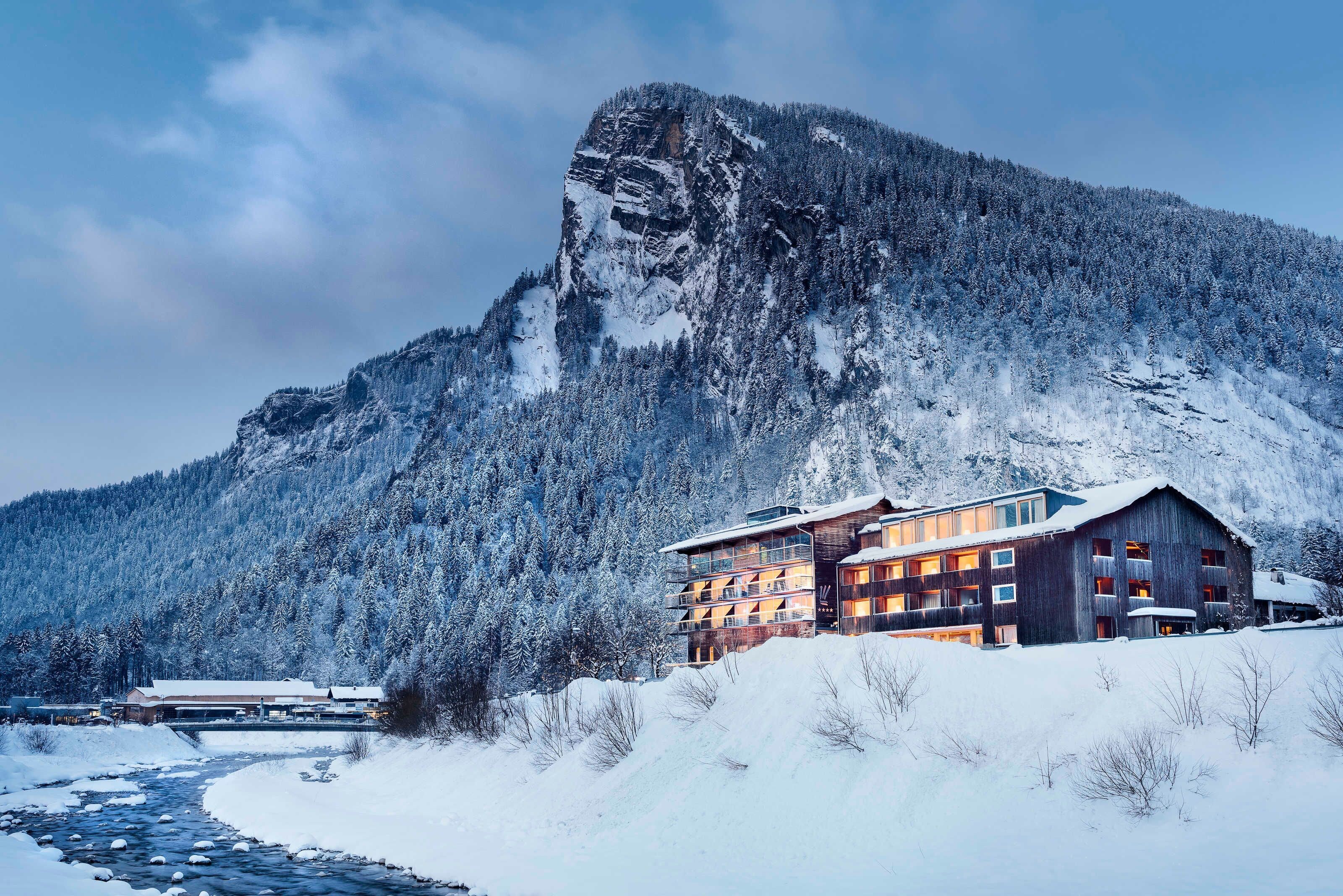 A mountain lodge with a river in front, surrounded by snow-covered trees.