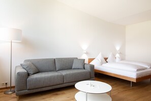 Une chambre moderne avec un canapé gris, une table ronde blanche, un plancher en bois et un lit avec une literie blanche.