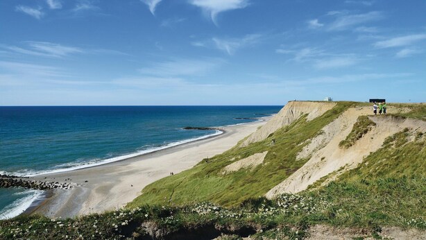 A coastal landscape with a sandy beach, grassy cliffs, and a clear blue sky.