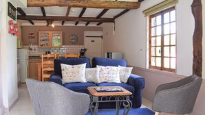 Un salon chaleureux avec un canapé bleu, deux fauteuils gris et une petite table basse en bois. On aperçoit une cuisine à l’arrière-plan, avec des meubles en bois et un pare-feu en carrelage.