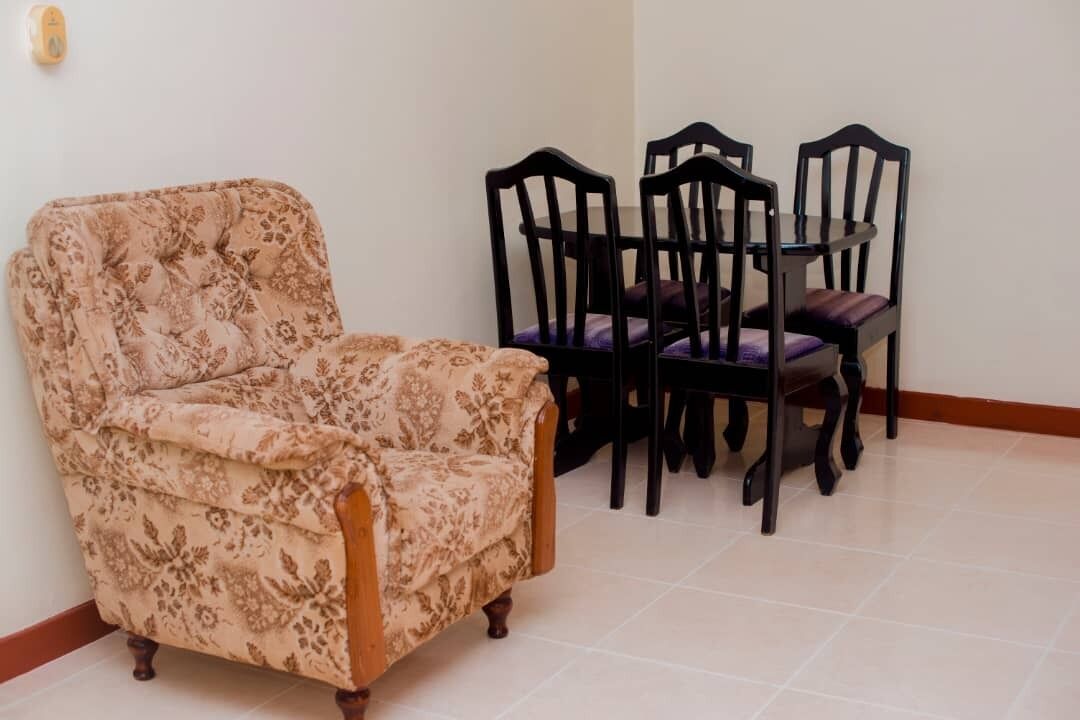 A room with a floral-patterned armchair, a round dining table with four chairs, and a tiled floor.