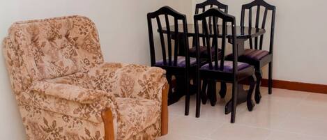 A room with a floral-patterned armchair, a round dining table with four chairs, and a tiled floor.