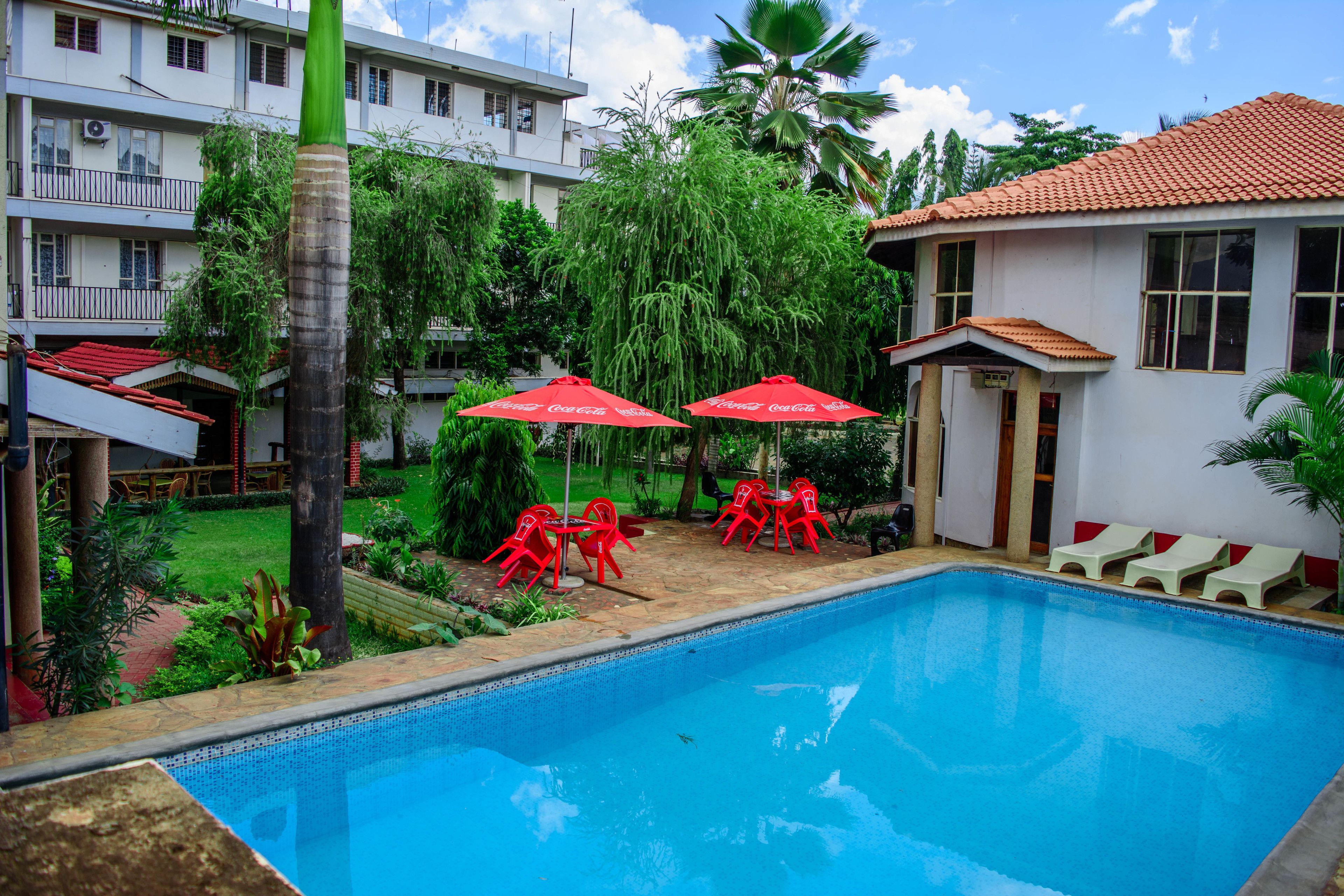 A swimming pool with red chairs and umbrellas, surrounded by greenery and a building with a red roof.
