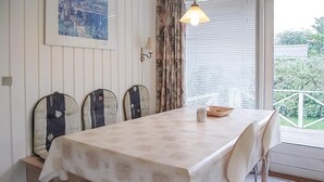 A dining area with a table covered in a floral tablecloth, four upholstered chairs, a hanging light fixture, a window with blinds, and a view of greenery outside. - 2 bedroom nice home in Hejls (Hejls)