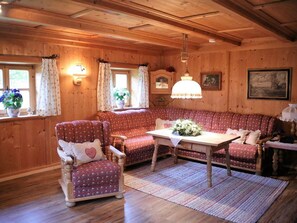 L’intérieur chaleureux d’un chalet, avec un revêtement mural en bois, un canapé rouge à motifs, un fauteuil à bascule en bois, une table agrémentée d’un centre de table floral et un luminaire suspendu.