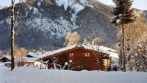 Eine Holzhütte mit schneebedecktem Dach, umgeben von schneebedeckten Bäumen und Bergen im Hintergrund.