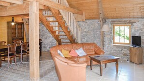 A cozy living room with a leather sofa, a wooden coffee table, and a stone wall. A wooden staircase leads to an upper level, and a dining area is visible in the background.
