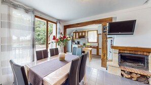 Een eetgedeelte met een tafel, stoelen en een bloevas. Achtergrond is een keuken met een televisie boven de open haard.
