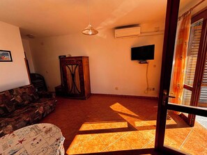 Uma sala de estar com sofá, um banquinho estampado, um armário de madeira e uma televisão na parede.