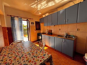 Uma cozinha com armários azuis, uma mesa de jantar com toalha de mesa florida e vista para o exterior através de uma porta.