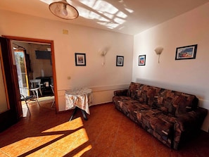 A living room with a patterned sofa, a small table with a newspaper-covered tablecloth, and wall-mounted artwork. - Holiday Apartment with Balcony and View (Crikvenica)