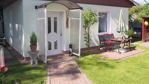 Een huis met een witte gevel, een rood dak en een deur met ruitpanelen. Buiten staat een bankje en een tafel met een parasol.