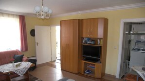 Een woonkamer met een bank, een tafel met kantkleed, een houten kast met televisie, en een keukenhoek met een koelkast.