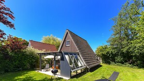 Une maison moderne avec un toit en pente, une véranda en verre et une pelouse agrémentée d’un fauteuil de jardin.