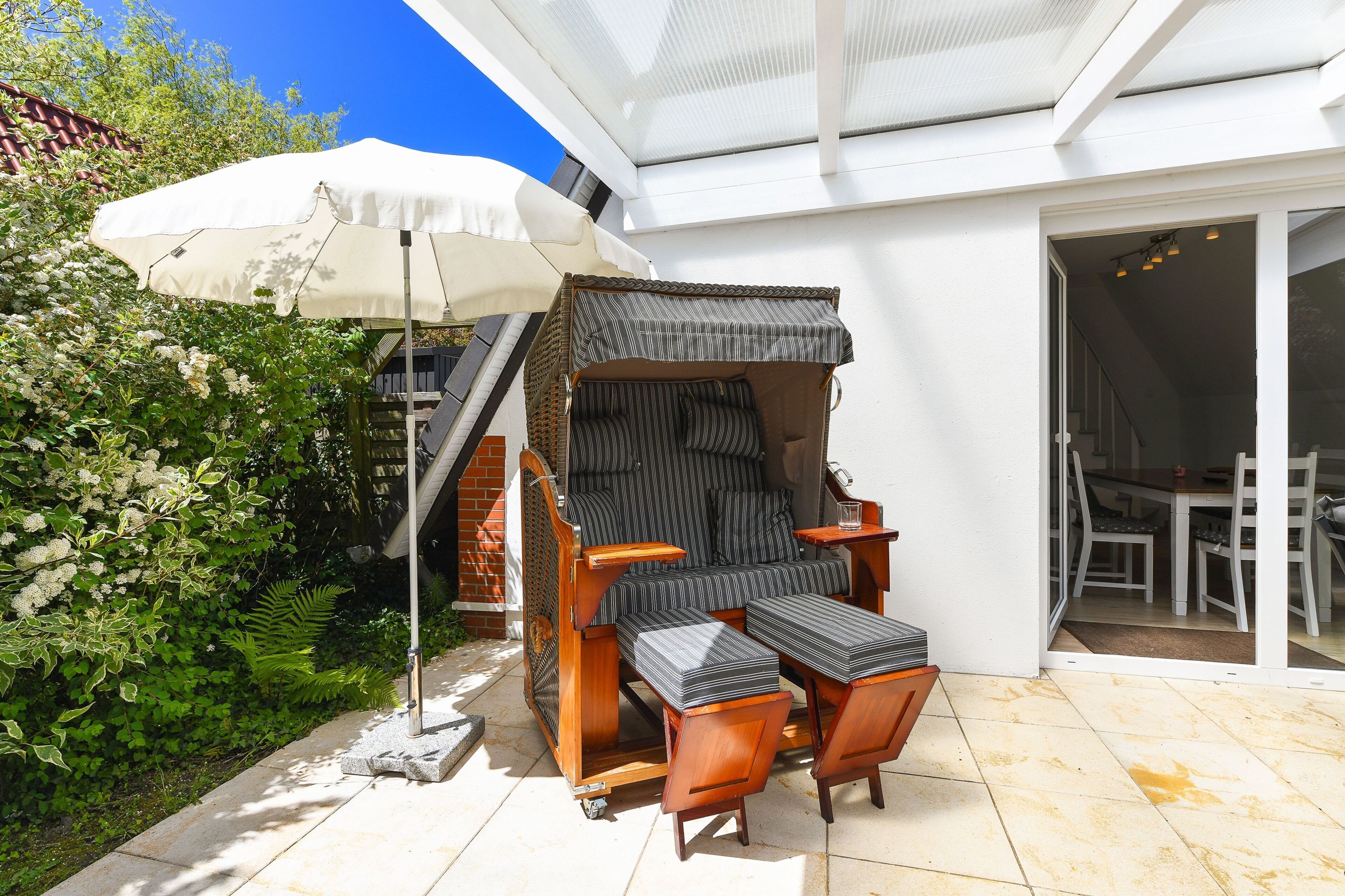 Eine Terrasse mit gepolstertem Stuhl und Hocker, einem weißen Sonnenschirm und einer Glastür, die zu einem Innenbereich führt.