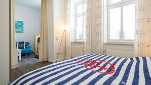 A bedroom with a bed, a standing lamp, a window with curtains, a blue chair, and a door leading to another room. - 24-08 - resort Villa Apt 24-08. (Kühlungsborn)