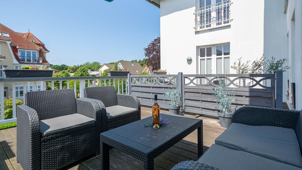 An outdoor patio with wicker furniture, a table with a bottle, and a white building in the background. - 24-08 - resort Villa Apt 24-08. (Kühlungsborn)