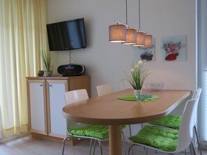 A dining area with a wooden table, green cushioned chairs, a flat-screen TV mounted on a cabinet, and a modern pendant light fixture.