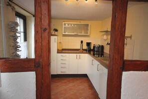 Una cocina con armarios blancos, estructura de vigas de madera, un ventanal con cortina y piso de baldosas.