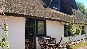 Une maison avec un toit de chaume, une porte-fenêtre coulissante et une terrasse en bois avec des chaises.