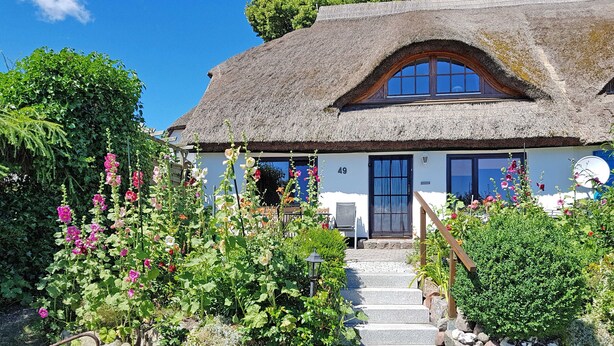 Una casa tradicional con techo de paja y una escalera de piedra que lleva a la entrada, rodeada de exuberantes jardines y plantas florecientes.