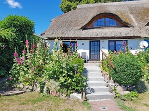 A traditional thatched-roof house with a stone staircase leading to the entrance, surrounded by lush gardens and flowering plants.