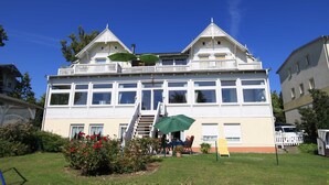 Un edificio grande de dos pisos con un balcón, una escalera y un pequeño jardín con una mesa y sillas.