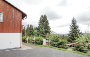 A wooden building with a gravel driveway leading to a fenced area with trees and a red-roofed structure in the background.