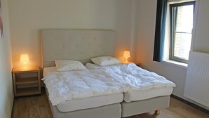 A double bed with a tufted headboard, two bedside tables with lamps, a window with a view, and wooden flooring. - Cozy apartment in Diksmuide with WiFi (Diksmuide)
