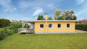 Ein eingeschossiges gelbes Gebäude mit Flachdach, Schornstein und einer kleinen Veranda mit einer Bank.
