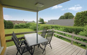 Een overdekt terras buiten met een tafel en stoelen, omgeven door een houten terras en uitzicht op een woongebied met huizen en groen.