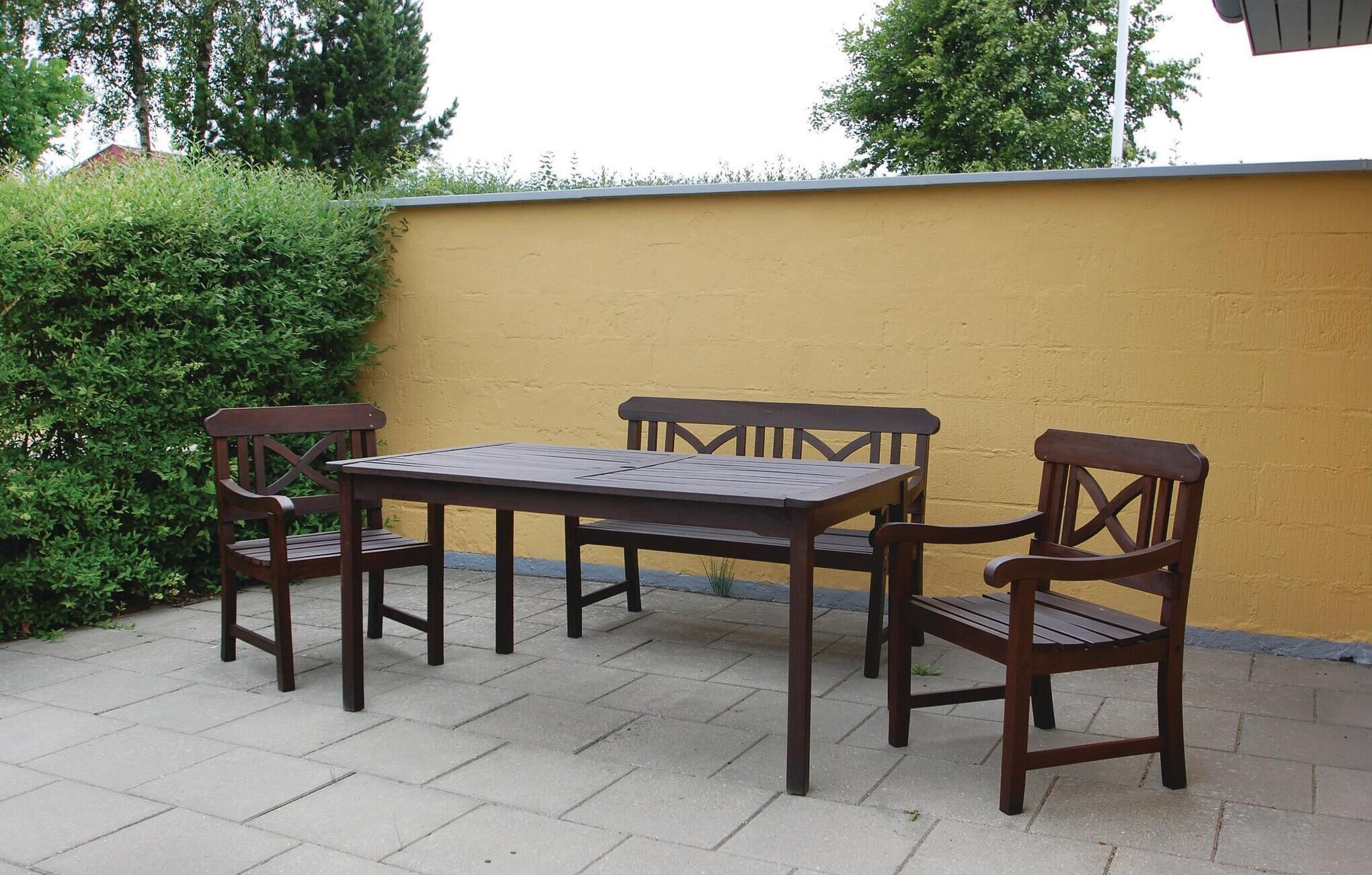 A patio with a wooden table and four chairs, a yellow wall, and green foliage.