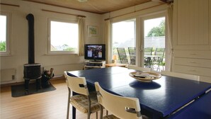 Ein gemütliches Zimmer mit Holzofen, einem Esstisch mit Stühlen, einem Fernseher und großen Fenstern mit Blick ins Freie.