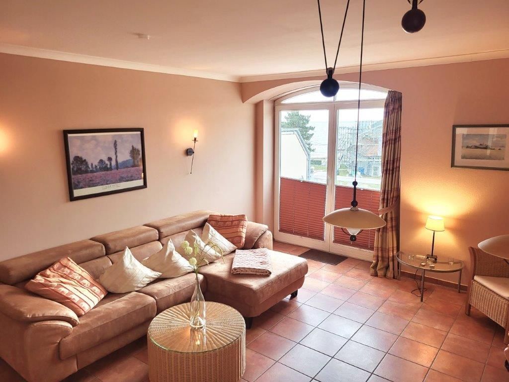 A living room with a sofa, a round wicker table, and a glass coffee table. There is a window with curtains, a lamp on a side table, and framed pictures on the walls.