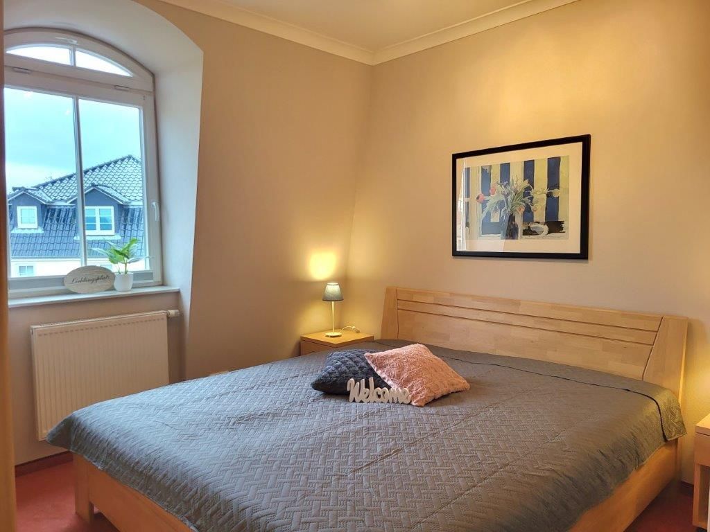 Ein Schlafzimmer mit Bett, Fenster, Nachttisch, Lampe und einem gerahmten Bild an der Wand.
