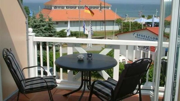 A balcony with two chairs and a table, overlooking a building with a red roof and flags.