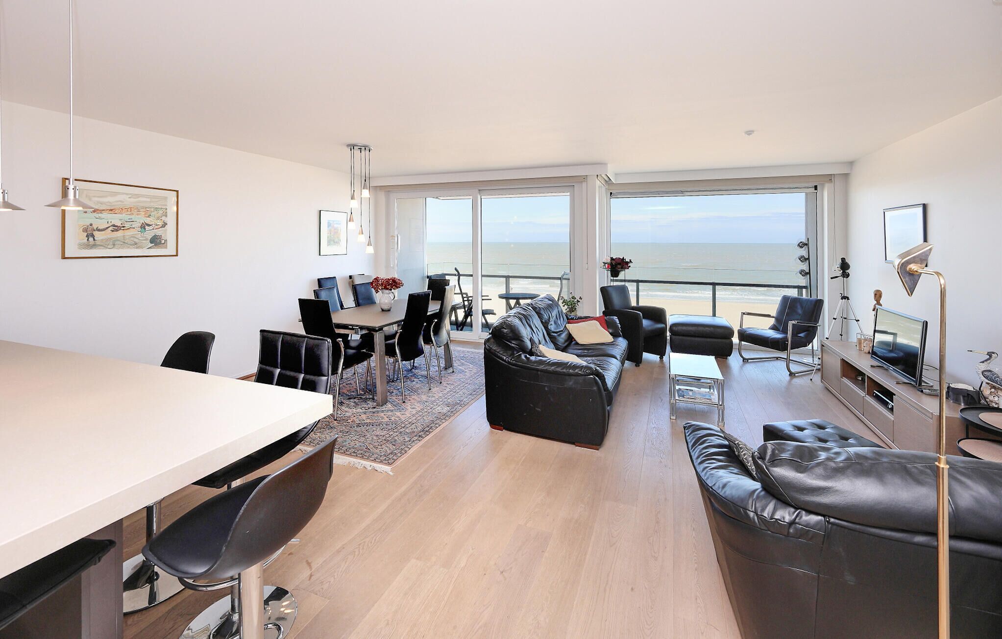 Ein modernes Wohnzimmer mit Essbereich, großen Fenstern mit Meerblick und einem Balkon mit Fernseher.