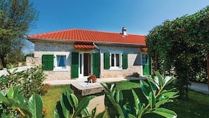 A stone house with a red-tiled roof, green shutters, and a small garden with plants and a flower pot.